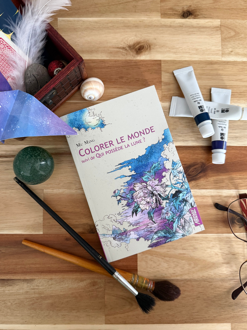 Livre Colorer le monde. Suivi de Qui possède la Lune? pris en photo avec à coté, des tubes de peintures, pinceaux, lunettes et une boite pleine de « trésors »: coquillage, origami de grue, plume, carte, caillou, boulet, etc.