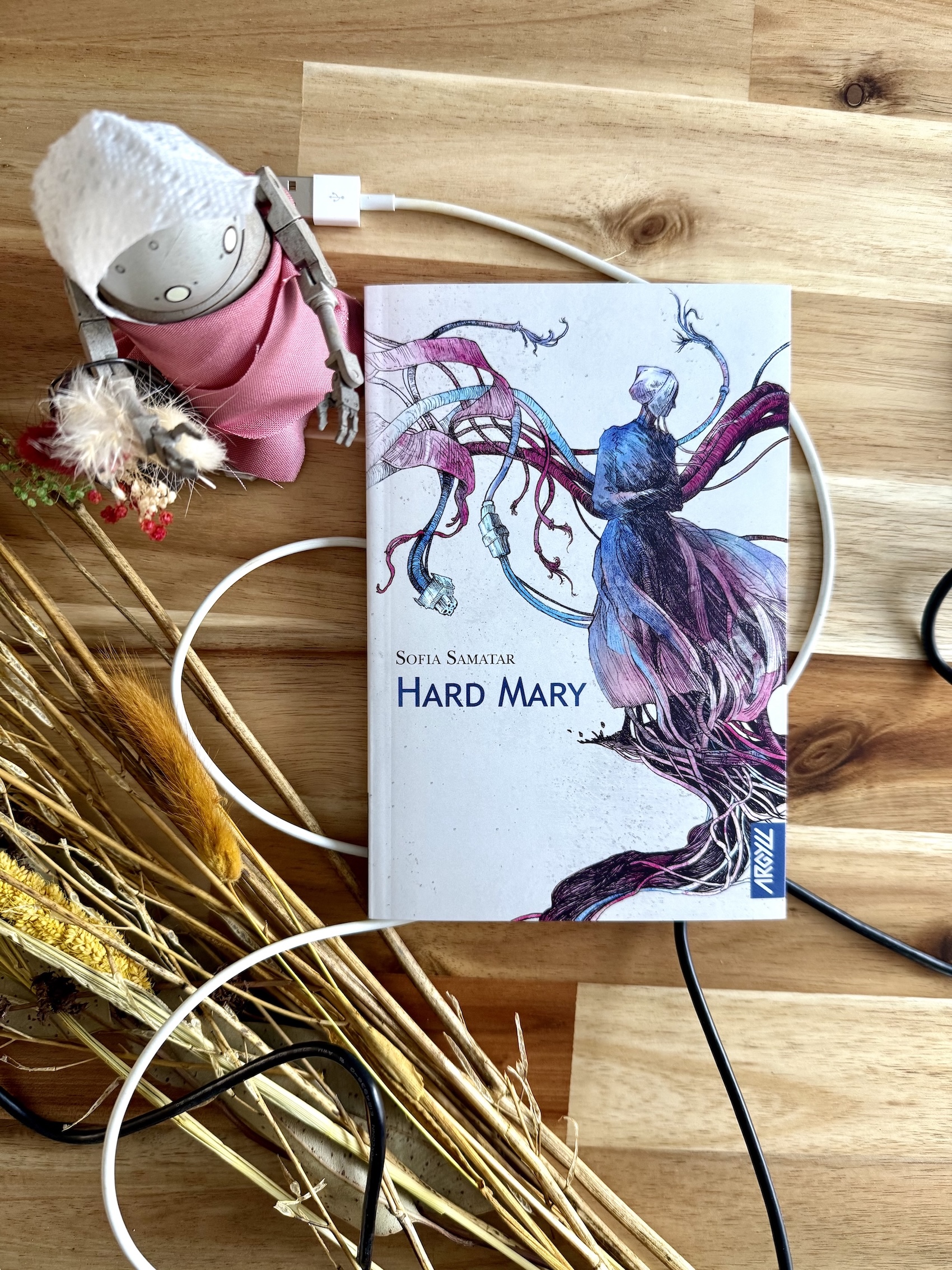 Livre posé à côté de fleurs et herbes séchées ainsi que des câbles. Est également posé à côté un robot habillé d'une robe et d'un bonnet blanc type mormon, avec des fleurs dans un seau pendu à son bras. La couverture d'Hard Mary montre une femme robot en tenu mormone depuis laquelle de nombreux câbles sortent.