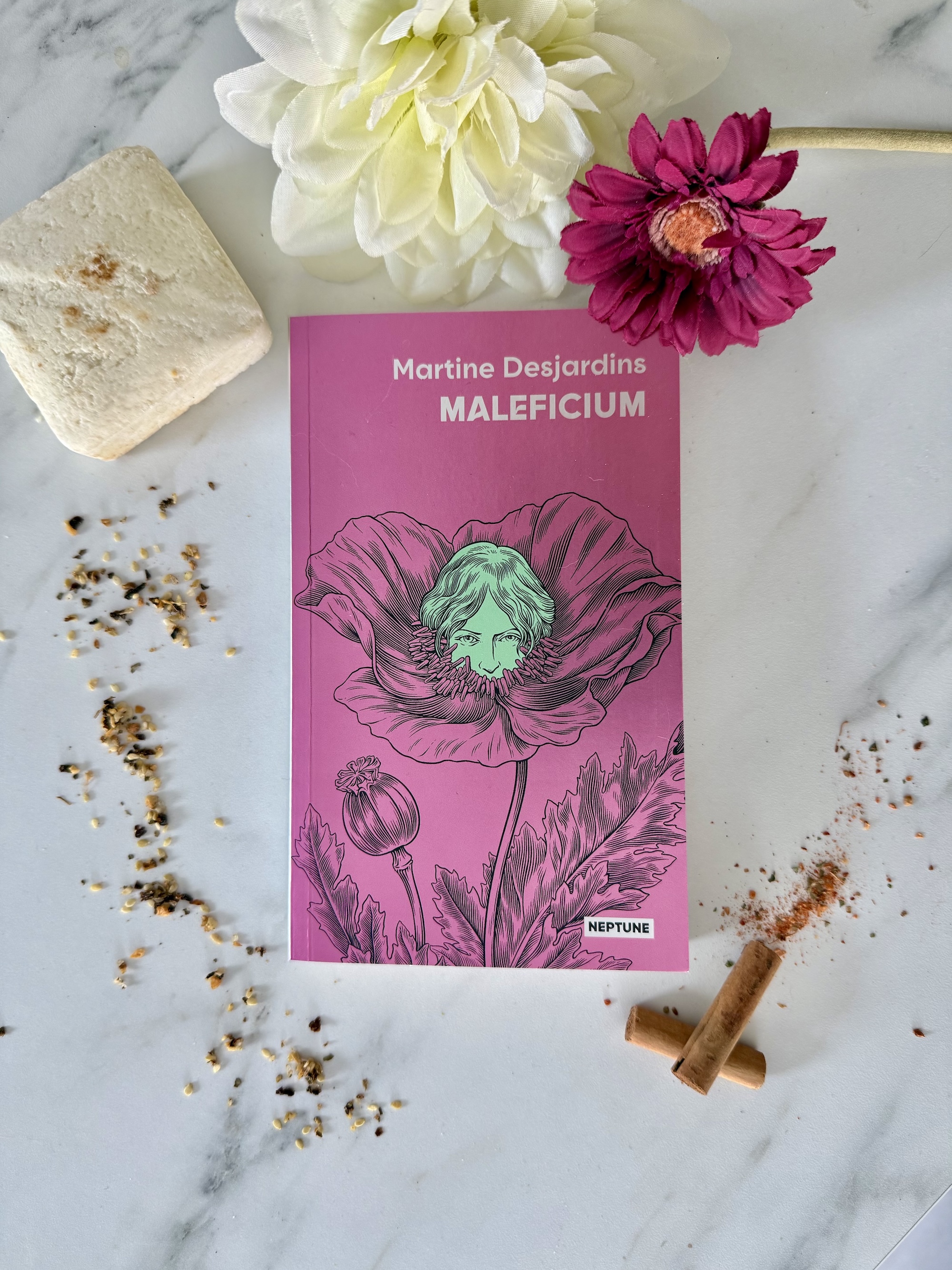 Livre posé à côté d’épices, cannelle, d’un savon artisanal et de fleurs mauves et blanches. La couverture de Maleficium montre un visage féminin émergeant du coeur d’une fleur. 