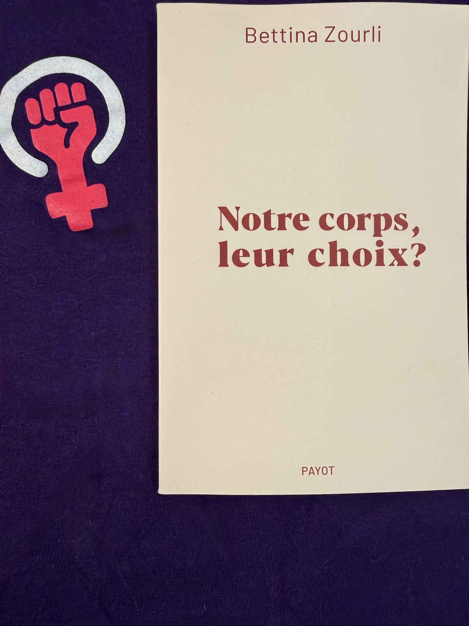 Livre posé sur un tissu mauve avec symbole féministe. La couverture de Notre corps, leur choix? montre simplement le titre et le nom de l'autrice en rouge sur fond beige