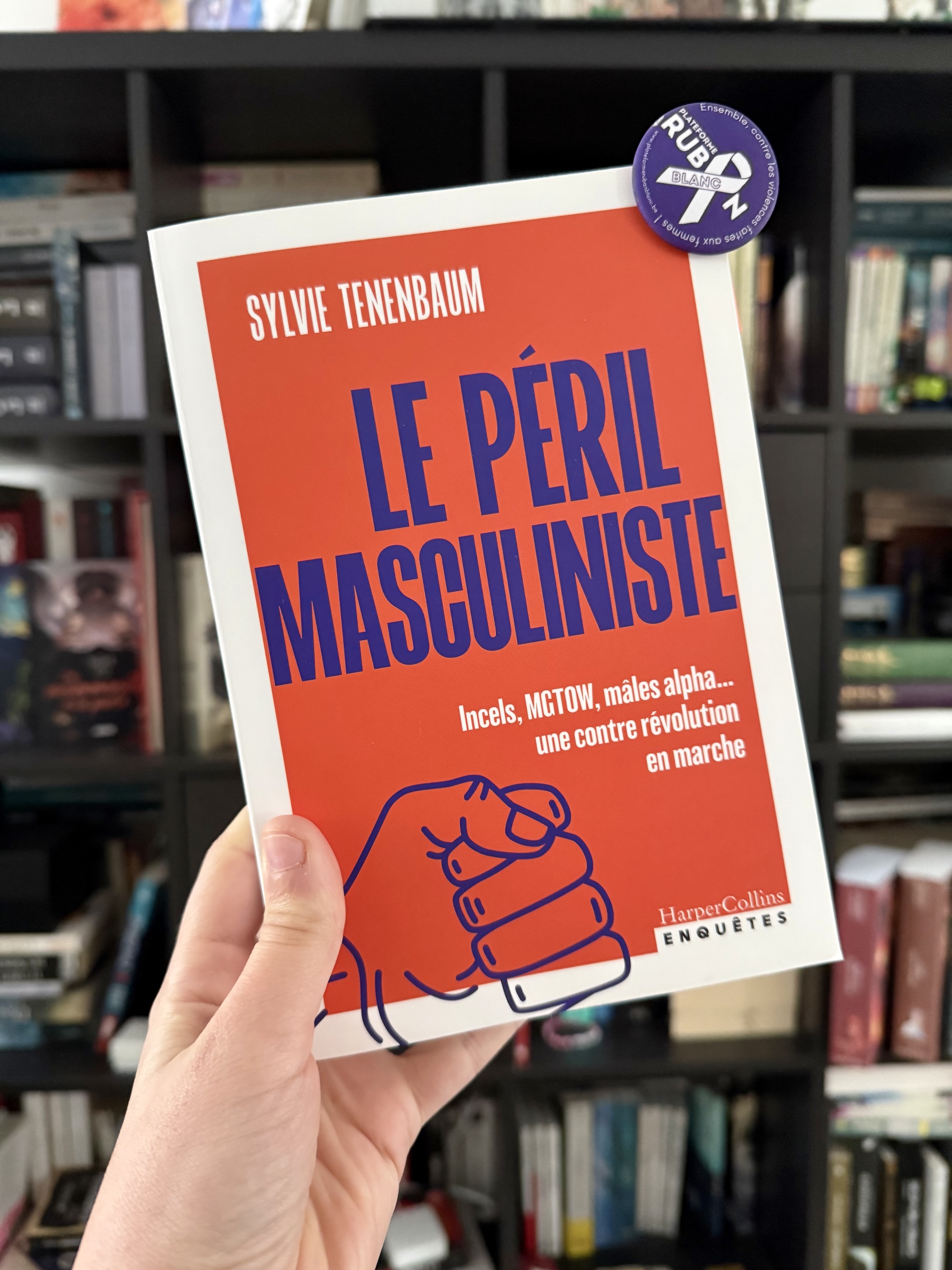 Livre tenu en main devant une bibliothèque. Au livre est accroché un badge de la Plateforme Ruban Blanc pour. la lutte contre les violences faites aux femmes. La couverture du Péril masculiniste montre le titre sur fond orange et un poing fermé de résistance.