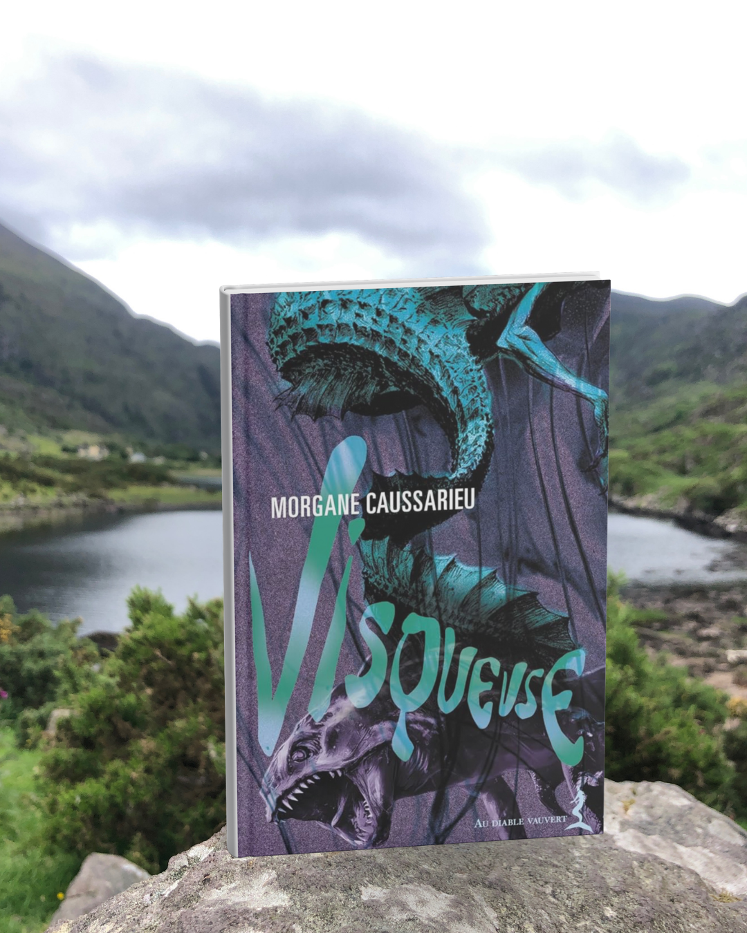 Livre pris en photo devant un lac entre deux montagnes. La couverture de Visqueuse montre une queue serpentine avec un torse humain et un poisson à grandes dents.