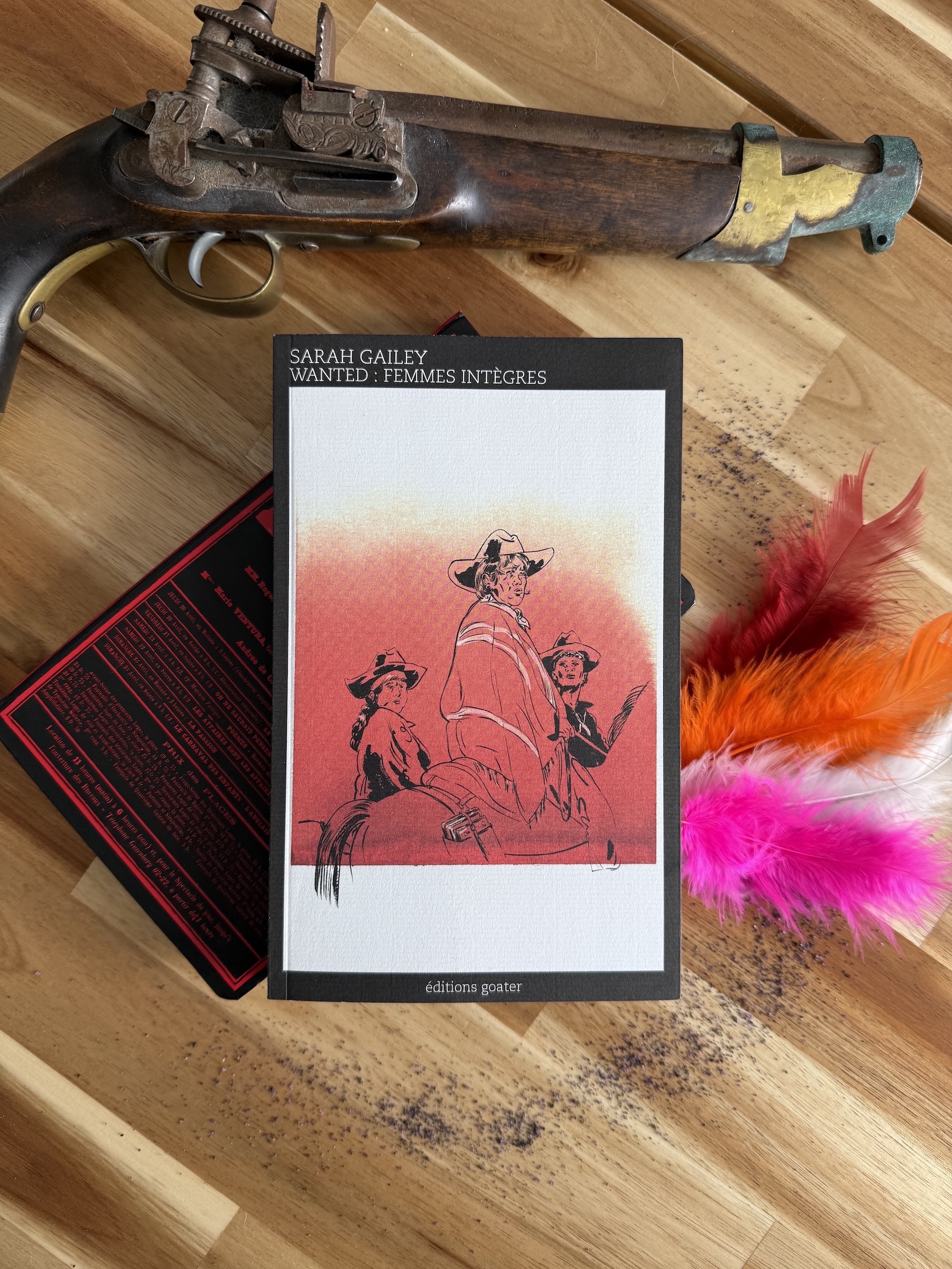  femmes intègres posé à côté d’un vieux fusil, d’un carnet de notes, de sable et de plumes aux couleurs du drapeau lesbien